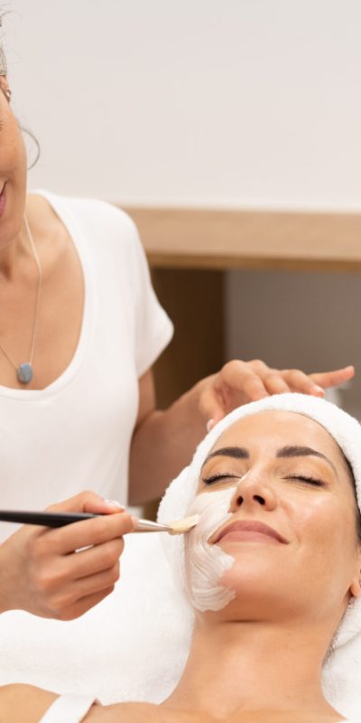 Smiling female cosmetologist applying hydrating mask with brush on face of female customer lying on table in beauty salon